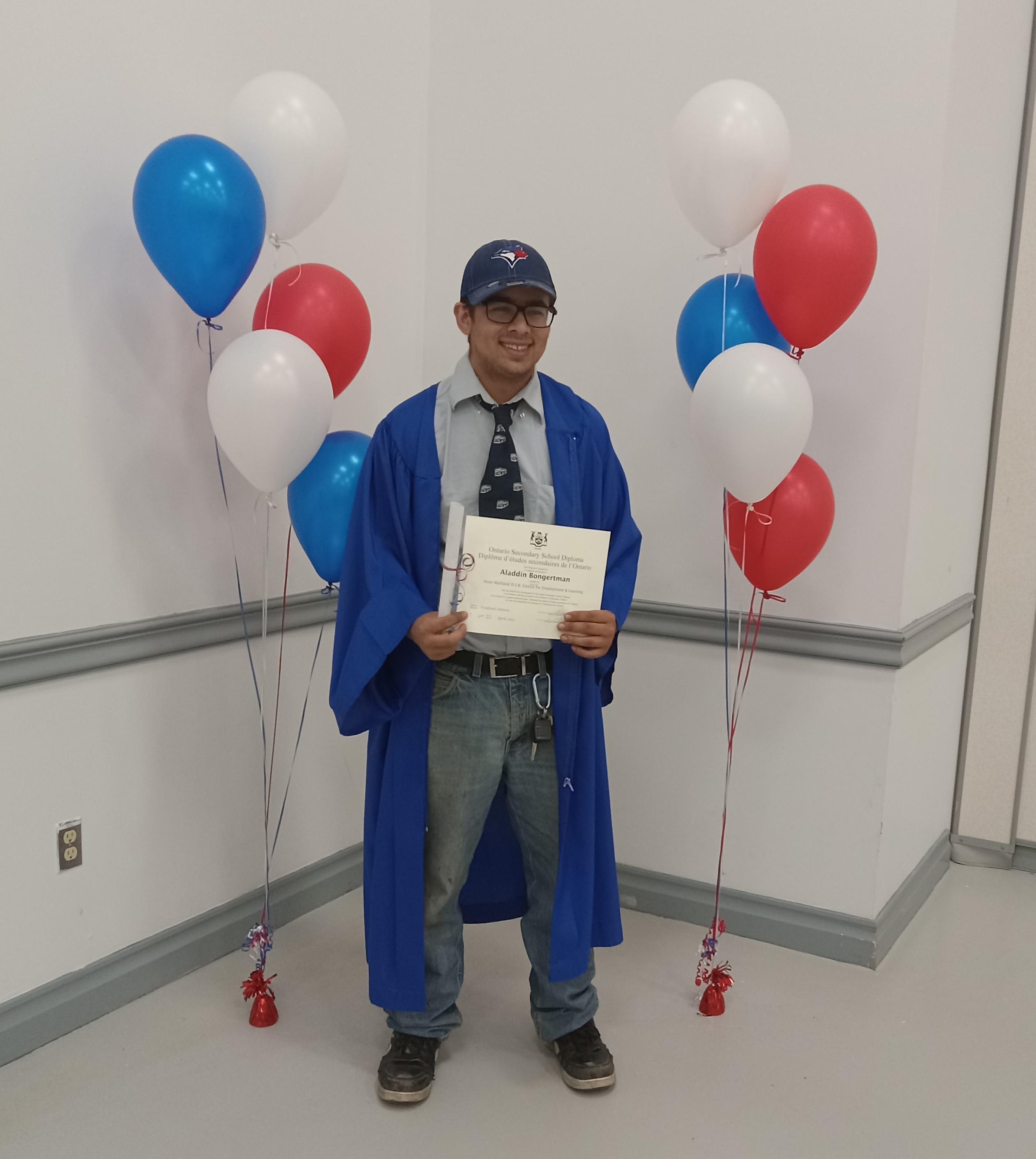 Aladdin poses in his gown with his diploma, red, white, and blue balloons in backgroundf